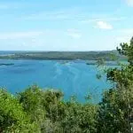 Ein malerischer Blick auf eine riesige blaue Lagune, umgeben von üppigem Grün. In der Ferne erstreckt sich ein schmaler Landstreifen ins Wasser, unter einem klaren blauen Himmel mit vereinzelten Wolken. Waldgebiete säumen den Horizont und schaffen eine friedliche, idyllische Landschaft.
