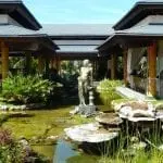 Eine ruhige Gartenszene mit einer steinernen Frauenstatue im Zentrum, umgeben von einem Koiteich. Offene Holzpavillons mit weißen Vorhängen flankieren den Teich. Üppiges Grün, darunter verschiedene Pflanzen und Sträucher, prägt die Landschaft unter einem strahlend blauen Himmel.