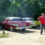 Ein Mann im roten T-Shirt steht neben zwei Oldtimern – perfekt für einen Familienausflug. Die rot-weißen und gelb-weißen Fahrzeuge stehen auf einem Feldweg, gesäumt von üppig grünen Bäumen. Sonnenlicht fällt durch die Äste und wirft nostalgische Schatten, die an den Stil der 1950er Jahre erinnern.