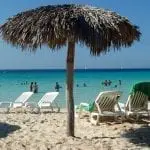 Ein Sandstrand, ideal für einen Familienurlaub, mit einem Strohschirm und mehreren weißen Liegestühlen, einer davon mit einem grünen Handtuch geschmückt. Familien genießen das Schwimmen und Waten im klaren türkisfarbenen Meer unter dem strahlend blauen Himmel.