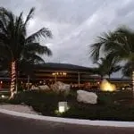 Der beleuchtete Eingang des tropischen Resorts begrüßt die Gäste in der Abenddämmerung mit zwei Palmen, die mit Lichterketten geschmückt sind und den Weg säumen. Im Vordergrund befindet sich ein angelegter Garten mit Sträuchern und großen Felsen, ideal für einen Familienausflug. Der bewölkte Himmel und das warme Licht laden Familien zu einer perfekten Familienzeit ein.