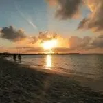 Ein atemberaubender Sonnenuntergang über einem Strand wirft goldene Strahlen über den Himmel – perfekt für „Urlaub mit Kind“. Die Sonne sinkt nahe dem Horizont unter und spiegelt sich in den sanften Wellen, während Familien ihre Familienzeit genießen und unter einem teilweise bewölkten Himmel am Sandstrand entlangschlendern.
