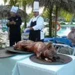 Köche mit weißen Hüten stehen unter einem Strohdach neben einem gebratenen Schwein auf Platten. Palmen und ein Swimmingpool mit einer Person in Badebekleidung, die auf einem Liegestuhl sitzt, sind zu sehen und suggerieren eine tropische Kulisse, perfekt für einen Familienurlaub.