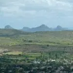 Eine weite Landschaft entfaltet sich, perfekt für einen Familienausflug, mit einer kleinen Stadt im Vordergrund, verstreuten Gebäuden und grünen Feldern. Sanfte Hügel erstrecken sich in den Mittelgrund, während einzigartig geformte Berge unter einem bewölkten Himmel aufragen.