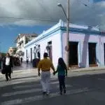 Eine Straßenszene entfaltet sich mit einem hellvioletten Gebäude, das an der Ecke durch blaue Verzierungen akzentuiert wird. Während eines Familienausflugs geht ein Mann in gelbem Hemd und weißer Hose Hand in Hand mit einer Frau in blaugrünem Top und Jeans. Andere Fußgänger schlendern in der Nähe unter dem teilweise bewölkten Himmel über dem Zebrastreifen.