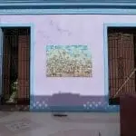 Eine hellviolette Wand mit zwei blau umrahmten, vergitterten Fenstern lädt zum Entdecken ein. Dazwischen entfaltet ein Wandgemälde eine Dorfszene – die perfekte Inspiration für einen Familienurlaub. Schatten eines Zauns tanzen über die Wand, und rechts lugt ein Teil eines roten Pflanzkübels mit grünen Pflanzen hervor.