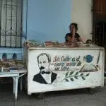 Eine Frau sitzt hinter einem weißen Bücherwagen – perfekt für einen Familienausflug. Auf dem Wagen ist ein gemaltes Männerporträt zu sehen und die Aufschrift „Ser culto es el único modo de ser libre“ (dt.: „Ser Kulto ist die einzige Art, frei zu sein“). Weitere Bücher liegen auf einem Tisch links. Die Außenkulisse besticht durch ein vergittertes Fenster und eine hellblaue Wand im Hintergrund.