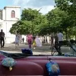 Im Vordergrund parkt ein mit „Happy Birthday“-Ballons geschmückter Oldtimer und sorgt für festliche Stimmung bei einem Familienausflug. Im Hintergrund schlendern Menschen über einen sonnenbeschienenen Platz, gesäumt von Bäumen und einem alten Gebäude. Eine Person in Pink, ähnlich einem Quinceañera-Kleid, sorgt unter dem leicht bewölkten Himmel für einen besonderen Charme.