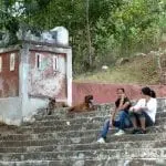 Zwei Frauen genießen ihre Familienzeit auf Steintreppen neben einem verwitterten rot-weißen Gebäude. Eine schlägt die Beine übereinander, die andere lehnt sich nach vorne, während zwei Hunde in der Nähe faulenzen. Die Umgebung ist ruhig, umgeben von Waldgebieten und dichtem Grün im Hintergrund.