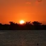 Sonnenuntergang über einem ruhigen See, perfekt für Familienzeit. Die Sonne steht tief am Horizont und wird teilweise von Bäumen in der Ferne verdeckt. Der Himmel ist in einem leuchtenden Orange- und Rotton gehalten und spiegelt sich im Wasser darunter. Die Baumsilhouetten bilden einen dunklen Umriss vor dem farbenfrohen Himmel – eine friedliche und ruhige Szene.