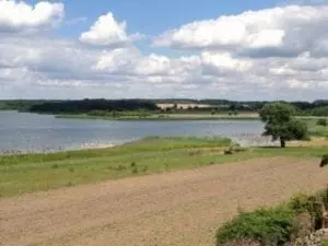 Ein malerischer Blick auf einen See unter bewölktem Himmel bietet die perfekte Kulisse für einen Familienausflug. Im Vordergrund ist ein gepflügtes Feld mit Grasflächen zu sehen, während im Hintergrund das ruhige Wasser des Sees zu sehen ist, umgeben von Grün und Bäumen in der Ferne – ein idealer Ort für einen Familienausflug.