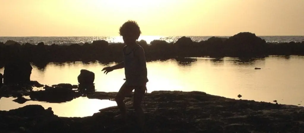 Silhouette einer Kind spielt während eines Familienurlaubs bei Sonnenuntergang an einem felsigen Ufer. Das Meer und die Sonne im Hintergrund schaffen eine ruhige und warme Atmosphäre, perfekt für einen Familienausflug.