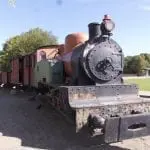 Eine historische Dampflokomotive mit großem Rundkessel und schwarzem Schornstein steht auf Gleisen und zieht rote und grüne Waggons. Perfekt für einen Familienausflug: Sie steht auf einer Wiese mit Bäumen und strahlend blauem Himmel und verspricht Abenteuer für alle an Bord.