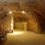 Ein rustikaler Keller mit Backsteinwänden und gewölbten Decken bietet die perfekte Kulisse für einen Familienausflug. Holzkisten sind ordentlich gestapelt, während ein kleiner Tisch zum gemütlichen Beisammensein einlädt, alles sanft beleuchtet von warmen Deckenleuchten.