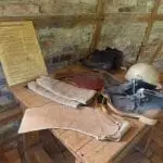 An einer Backsteinwand steht ein Holztisch, auf dem alte Arbeitskleidung wie Stiefel und Handschuhe ausgestellt sind – eine Erinnerung an einen Familienausflug in eine alte Fabrik. Ein Informationsplakat zum Thema Arbeitssicherheit steht auf dem Tisch und bietet Einblicke in die Vergangenheit.