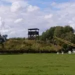 Eine Wiese mit einem rustikalen Wachturm auf einem kleinen Hügel in der Ferne ist ideal für einen Familienausflug. In der Nähe stehen Baumaschinen bereit, darunter ein blauer Kran. Bäume umgeben den Hügel, und der Himmel ist teilweise bewölkt, was den Charme dieser friedlichen Szenerie noch verstärkt.