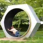 Ein Kind, wahrscheinlich auf einem Familienausflug, spielt in einer großen sechseckigen Metallkonstruktion auf dem Rasen. In beigefarbenem Hemd und Jeans vergnügt es sich unter dem sonnigen Himmel. Bäume und eine Bank verleihen der ruhigen Kulisse ihren Charme.