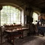 Eine rustikale Werkstatt mit Backsteinwänden und Holzwerkbänken voller Werkzeuge und Holzklötze – perfekt für einen Familienausflug. Große Bogenfenster lassen Tageslicht herein und geben den Blick auf den Wald frei. Neben einer der Bänke steht eine alte Bandsäge auf dem Steinboden.