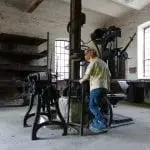 Ein junges Kind mit roten Haaren beschäftigt sich in einer alten Werkstatt mit antiken Maschinen. Der Raum, perfekt für einen Familienausflug, hat große Fenster, Steinwände und Regale. Das Kind, bekleidet mit einem beigefarbenen Hemd und blauen Jeans, steht neugierig neben den Metallgeräten.