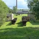 Eine Wiese mit zwei rostigen, ungenutzten Gleisen lädt zu einem Familienausflug zu einem entfernten Fabrikgebäude ein. Alte Eisenbahnwaggons stehen auf den Gleisen, eingerahmt von Bäumen und historischen Backsteingebäuden. Ein hoher Schornstein wacht unter dem bewölkten Himmel und lädt dazu ein, die Geschichten vergangener Zeiten zu erkunden.