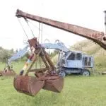 Ein rostiger alter Bagger mit einer großen Metallklaue, geschmückt mit einer bunten Strickhülle, steht auf einer Wiese – ein skurriler Anblick für jeden Familienausflug. Ein blaues Fahrzeug ist daran befestigt, im Hintergrund ist eine Holzkonstruktion zu sehen. Der Himmel ist teilweise bewölkt.