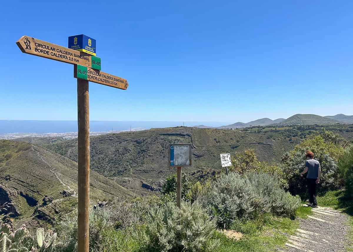 Eine Person steht in der Nähe eines Wanderwegs auf einem grünen Hügel, direkt neben einem hölzernen Wegweiser mit Wegbeschreibungen – eine einladende Szenerie, wie sie typisch für „Wandern mit Kindern auf Gran Canaria“ ist. Unter dem strahlend blauen Himmel sind in der Ferne Berge und eine Stadt zu sehen, die Familien Abenteuer und Entdeckungen versprechen.