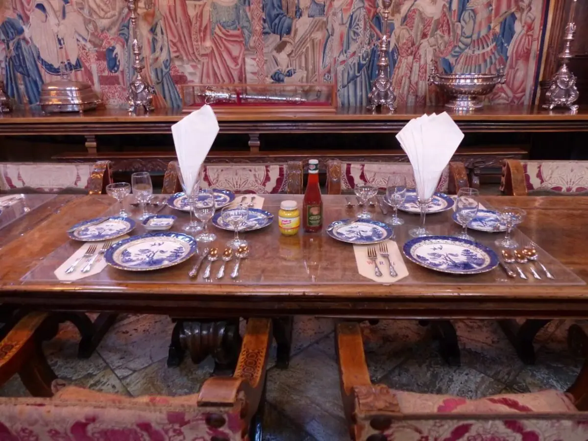 Ein Esstisch mit blau-weiß gemusterten Tellern, Gläsern und Besteck wartet auf ein Familienfestmahl. Servietten sind hochkant gefaltet, und Gewürze wie Senf und eine Soßenflasche zieren die Mitte. Hinter diesem charmanten Familientreffpunkt hängt ein dekorativer Wandteppich an der Wand.