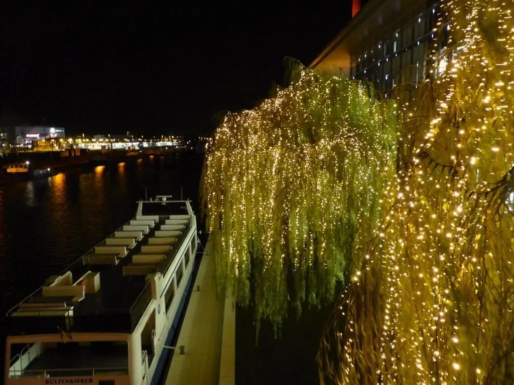 Ein angedocktes Boot liegt nachts neben einem Gebäude und einer Trauerweide, die mit leuchtenden Lichterketten geschmückt ist – perfekt für einen Familienurlaub. Die sanft beleuchtete Stadtlandschaft spiegelt sich im Wasser und schafft eine ruhige und festliche Atmosphäre, ideal für Familien.