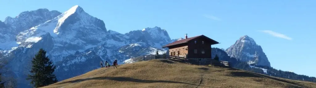 Ein kleines Holzhaus steht auf einem grasbewachsenen Hügel inmitten einer ruhigen Berglandschaft. Schneebedeckte Gipfel und ein strahlend blauer Himmel bilden die Kulisse. Eine Gruppe Wanderer, darunter ein neugieriger Junge, steigt den Hügel hinauf zum Haus – ein ideales Ziel für einen Familienausflug.