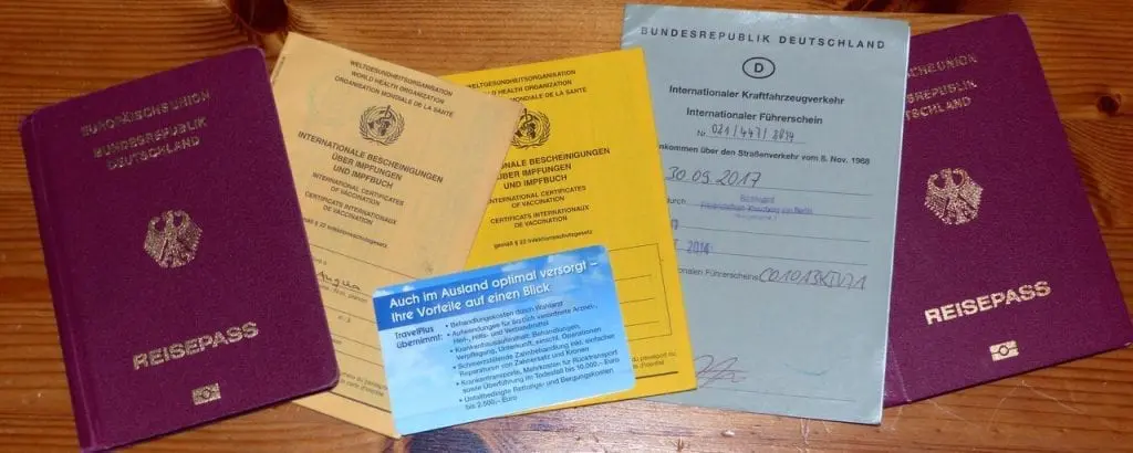 Vier Reisepasshefte, eine blaue Karte und zwei gelbe internationale Impfpässe liegen auf einer Holzfläche ausgebreitet, bereit für den Familienurlaub. Deutsche Texte und Embleme deuten auf die Abenteuer hin, die auf dieser Familienreise warten.