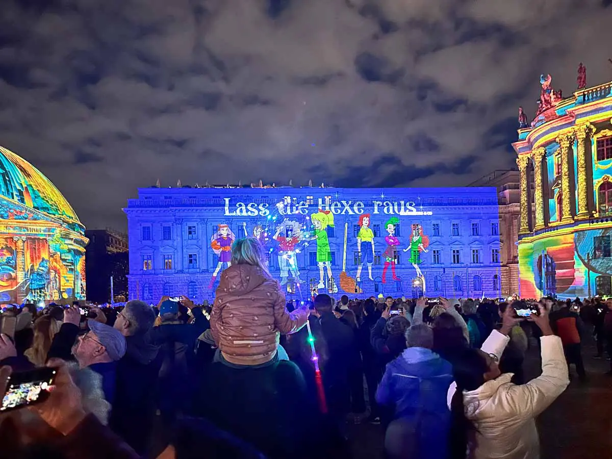 Eine Menschenmenge betrachtet nachts beim Festival of Lights eine farbenfrohe Lichtprojektion an einem Gebäude, bei der animierte Figuren und deutscher Text die Szene erhellen. Einige Leute machen Fotos, und in der Mitte sitzt ein Kind auf den Schultern eines Erwachsenen.