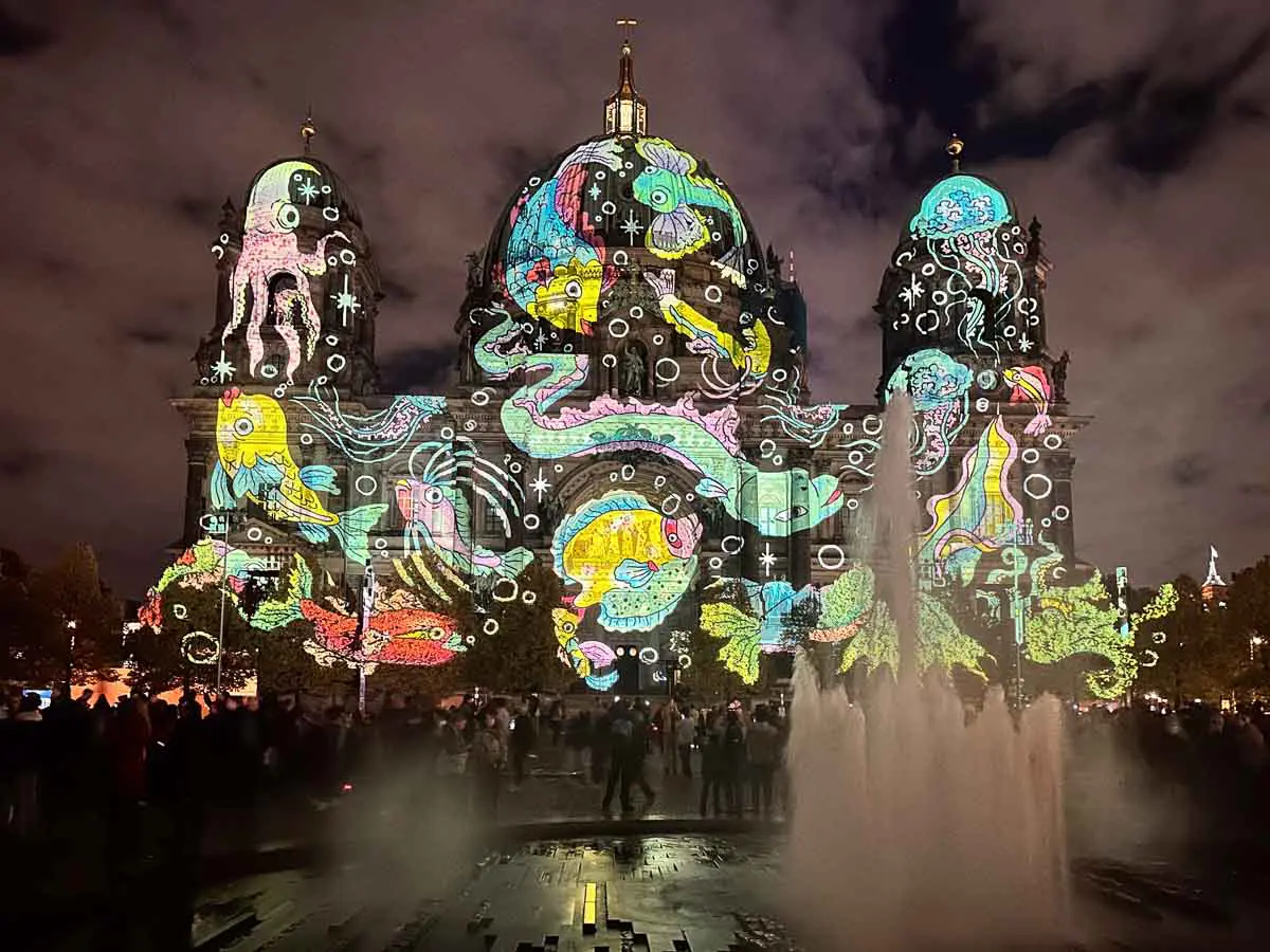 Die Fassade des Berliner Doms wird mit farbenfrohen Lichtprojektionen beleuchtet, die Meeresbewohner zeigen, während im Vordergrund ein Springbrunnen sprüht und sich nachts Menschen – darunter auch Familien, die das Lichterfest mit ihren Kindern genießen – versammeln, um das Spektakel zu beobachten.