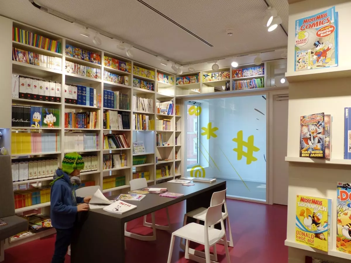 Ein Kind mit grüner Mütze und blauem Mantel liest an einem Tisch in einem lebhaften Raum, der an einen Familienurlaub im Fichtelgebirge erinnert. Die Regale sind randvoll mit Comics, und Poster von Helden schmücken die Wände. Ein roter Boden unter heller Beleuchtung trägt zur fröhlichen Atmosphäre bei.