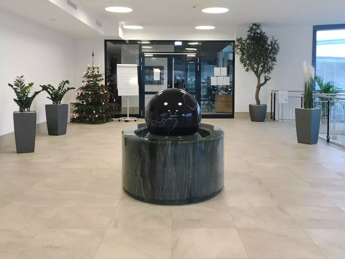 Die großzügige Lobby mit beigefarbenem Fliesenboden und einem zentralen schwarzen Kugelbrunnen lädt zum einladenden Start in Ihren Familienurlaub im Fichtelgebirge ein. Topfpflanzen, darunter ein geschmückter Weihnachtsbaum, zieren die Wände. Große Fenster und elegante Möbel schaffen eine moderne, luftige Atmosphäre.