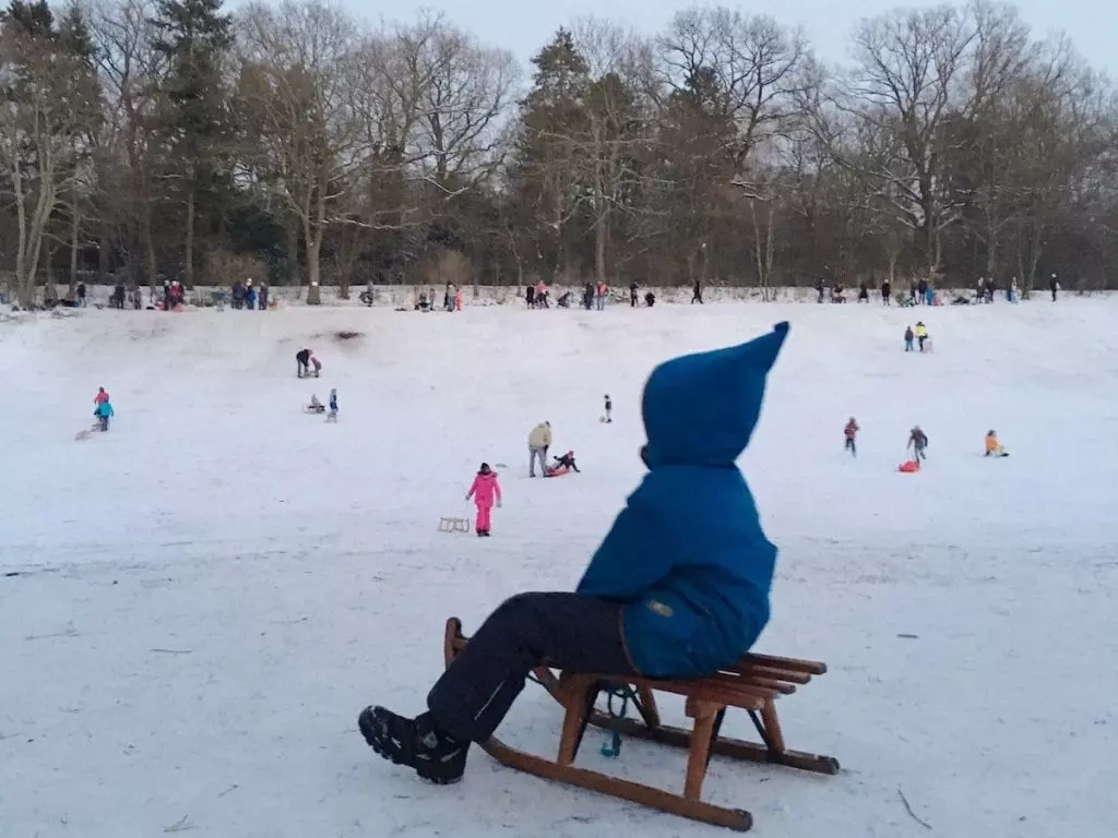 Eine Person in blauer Jacke und Kapuze sitzt auf einem Holzschlitten und beobachtet Familien, die den verschneiten Spielplatz genießen. Kinder und Erwachsene gleiten den sanften Hang hinunter und träumen dabei von zukünftigen Städtereisen mit Kindern in Berlin. Bäume säumen dieses Winterwunderland und runden die malerische Szenerie ab.