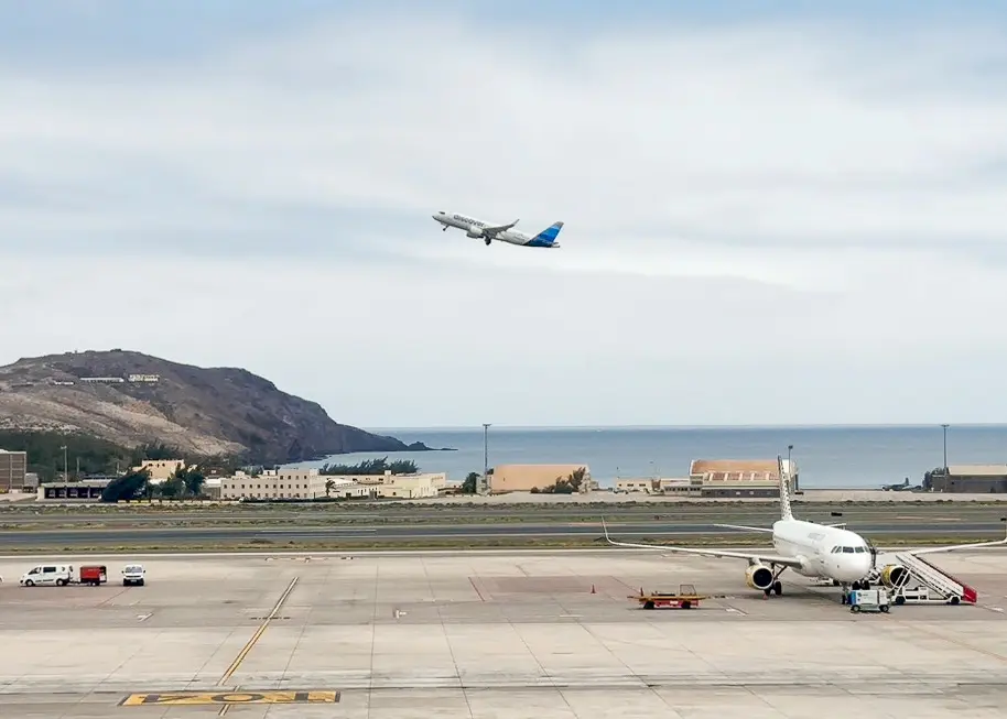Ein Flugzeug hebt über einer Start- und Landebahn in der Nähe des Meeres ab, während ein weiteres Flugzeug auf dem Vorfeld steht und mehrere Fahrzeuge in der Nähe zu sehen sind. Während Reisende günstige Flüge für die Ferien suchen, sind im Hintergrund unter einem bewölkten Himmel ein Hügel und einige Gebäude zu sehen.