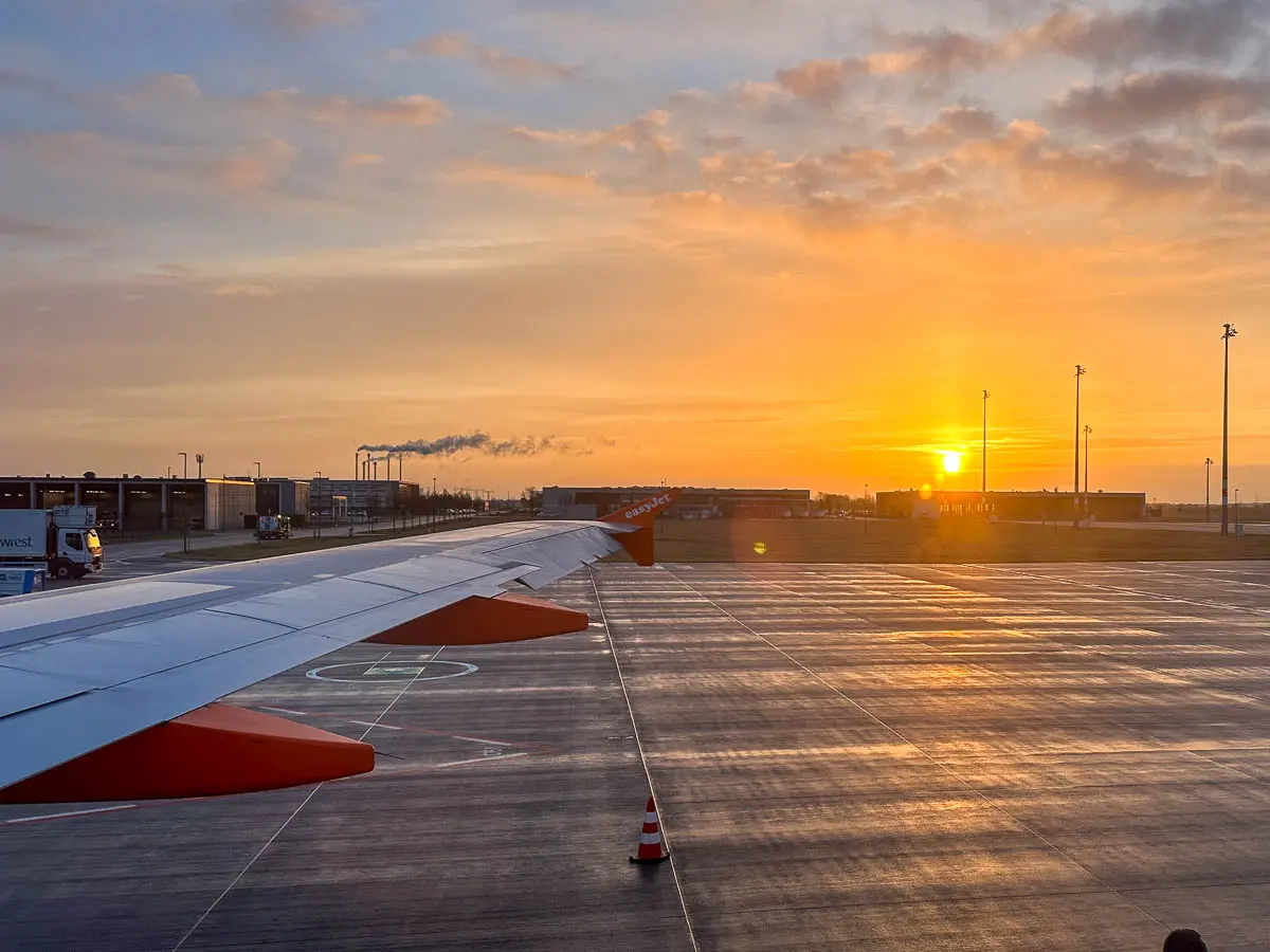 Die Sonne geht über einer Flughafenlandebahn unter, gesehen aus einem Flugzeugfenster. Der Flügel des Flugzeugs erstreckt sich über das gesamte Bild, und unter einem orangefarbenen Himmel sind Gebäude, Lichter und einige Fahrzeuge zu sehen – eine inspirierende Szene für alle, die  günstige Flüge für die Schulferien suchen.