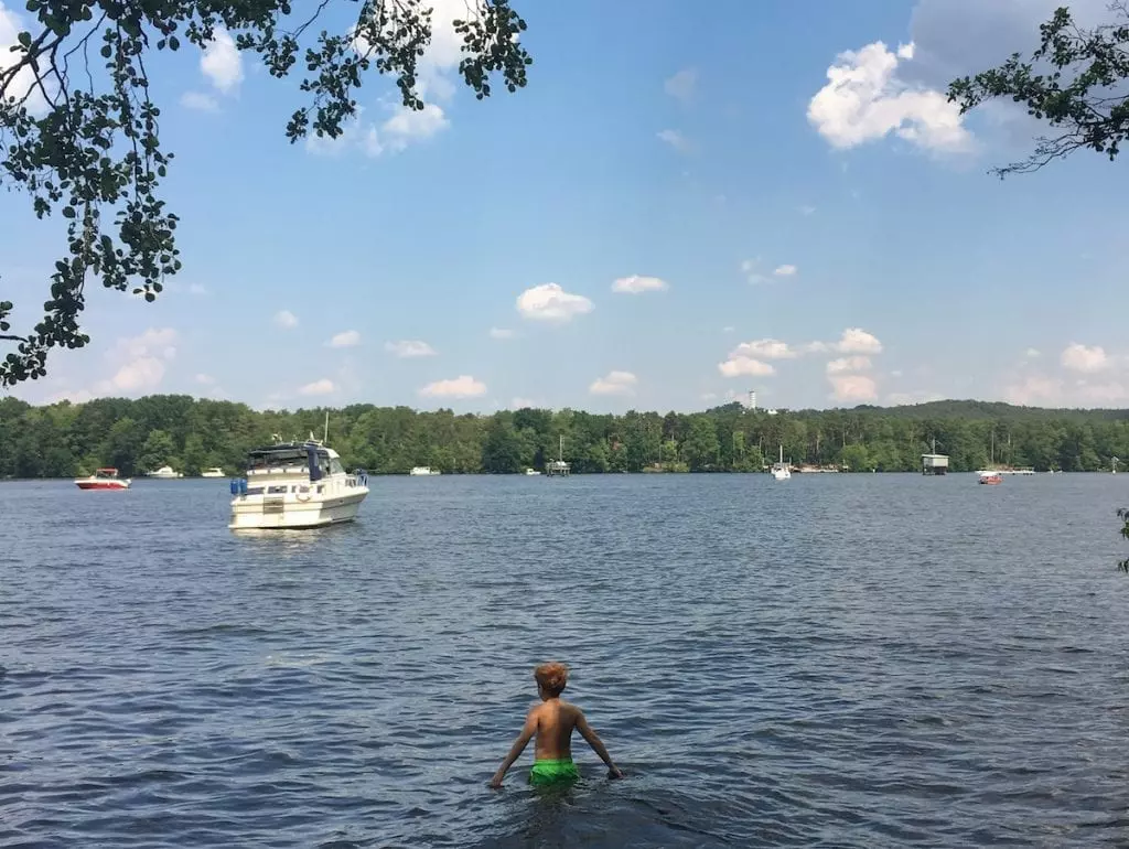 Ein Kind in grünen Shorts genießt das Baden in einem Berliner See, dessen Ufer von Bäumen gesäumt ist. Mehrere Boote treiben unter einem teilweise bewölkten Himmel.