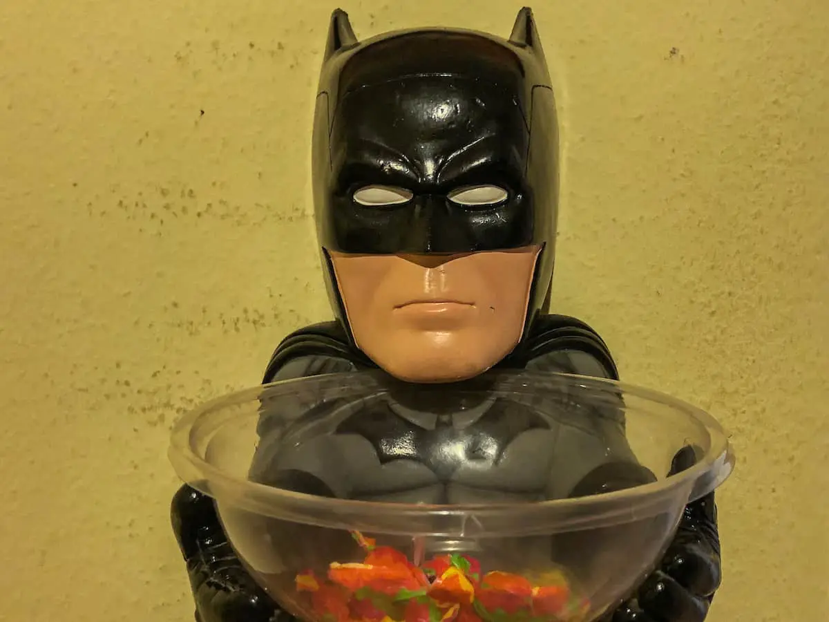 Eine Batman-Figur aus Kunststoff hält eine durchsichtige Schale mit bunten Bonbons in der Hand, vor dem Hintergrund einer schlichten beigen Wand – die perfekte Inspiration für dein nächstes Escape Game mit Kindern.