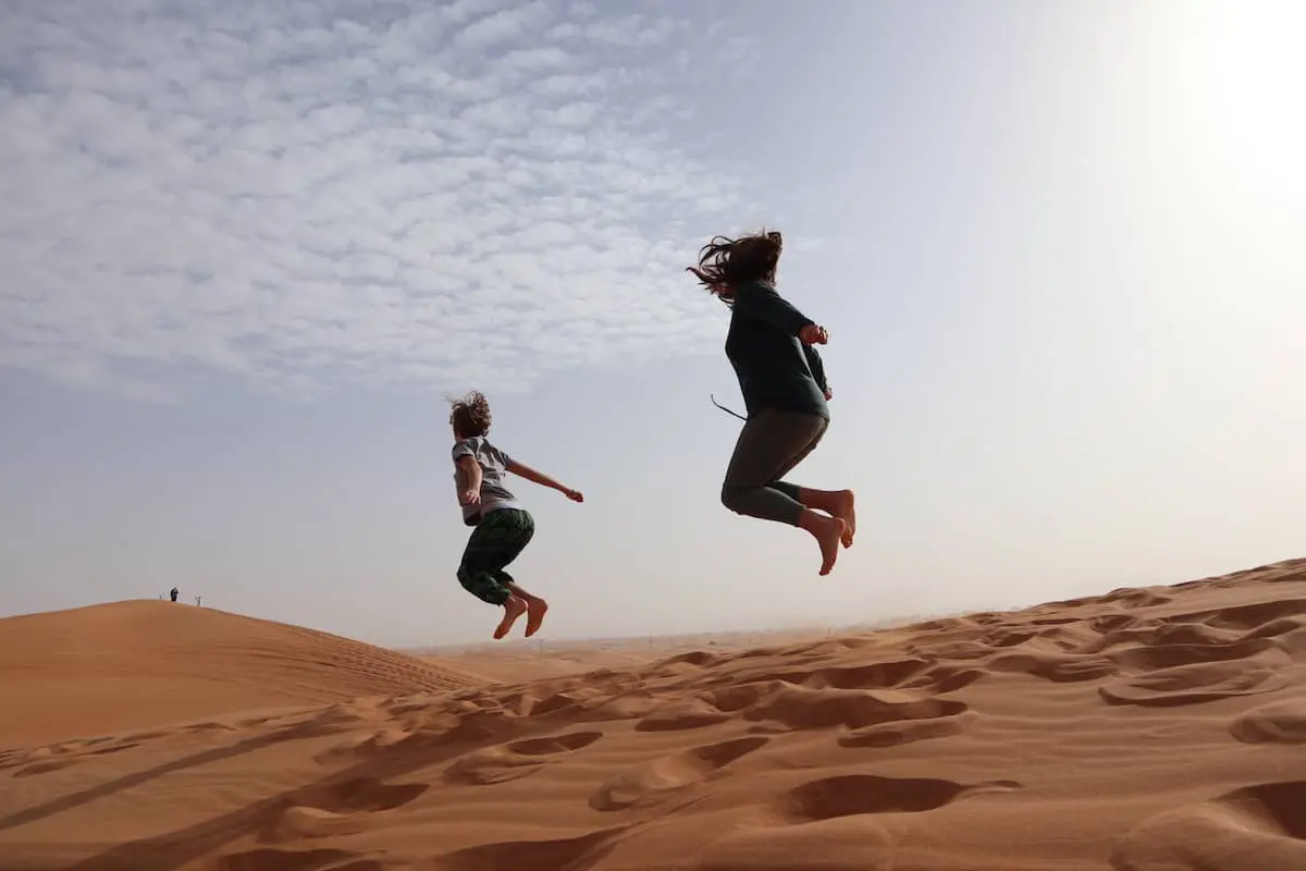 Zwei Menschen springen unter einem teilweise bewölkten Himmel auf Sanddünen in die Luft, ihre Fußspuren zeichnen sich im Sand ab – ein fröhlicher Moment aus dem Leben einer alleinerziehenden Familie auf Weltreise, während in der Ferne Menschen durch die goldene Landschaft schlendern.
