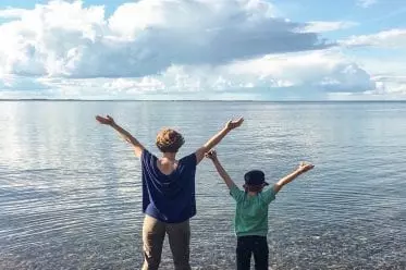 Mutter und Kind stehen mit ausgebreiteten Armen an einem klaren See in Sommerkleidung mit den Füßen im Wasser.
