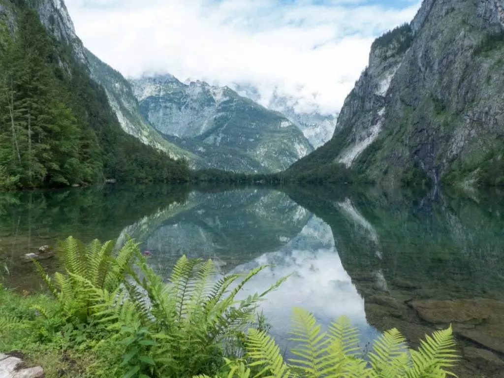 Ein ruhiger See in Berchtesgaden spiegelt die umliegenden Berge unter einem bewölkten Himmel wider. Üppige grüne Bäume und Farne rahmen den Vordergrund ein, während sich im Hintergrund hoch aufragende Gipfel mit Schneeflecken erheben. Diese Landschaft bildet eine friedliche Naturlandschaft, die zu den schönsten Sehenswürdigkeiten der Region zählt.