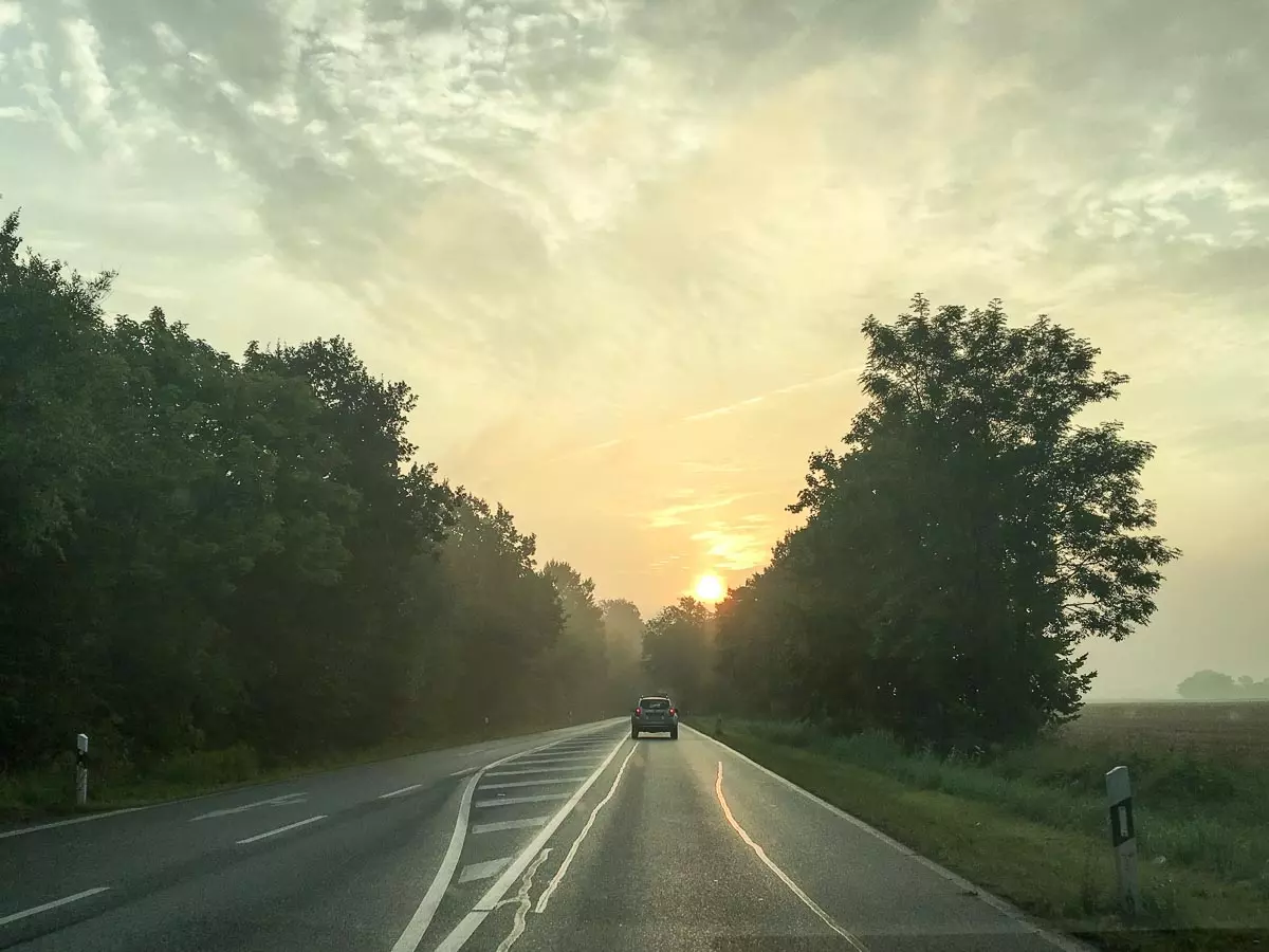 Ein Auto fährt auf einer Landstraße in Norddeutschland entlang, umgeben von Bäumen auf beiden Seiten. Die Sonne geht am Horizont auf und taucht den nebligen Himmel dieses ruhigen Roadtrips in einen warmen Schein. Der nasse Asphalt spiegelt das sanfte Morgenlicht.