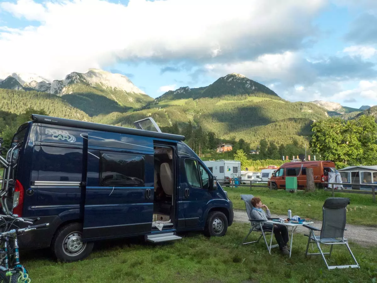 Ein blaues Wohnmobil steht auf einem malerischen Campingplatz im Herzen des Südens, mit Bergen im Hintergrund. Eine Person entspannt sich mit Getränken auf einem Stuhl an einem kleinen Tisch unter leicht bewölktem Himmel – perfekt für einen unvergesslichen Roadtrip. Campingausrüstung und Fahrräder stehen in der Nähe.