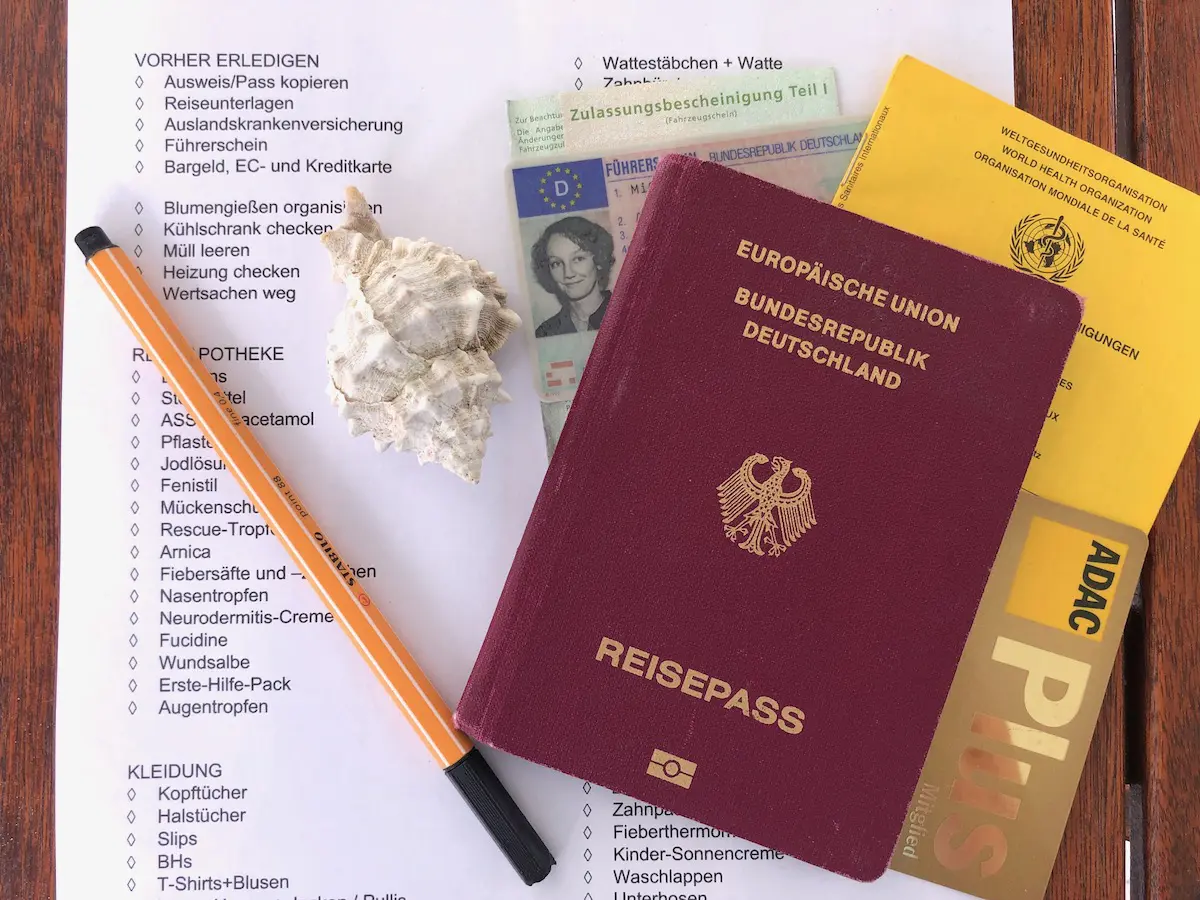 Ein deutscher Reisepass, ein ADAC-Heft und eine Muschel liegen auf einem Dokument mit der Aufschrift „Wohnmobil-Packliste“. Daneben liegt ein schwarz-oranger Stift auf dem Papier. Unter der Muschel lugt ein kleines Passfoto hervor.