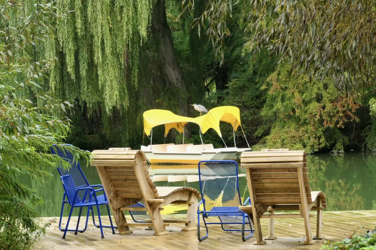 Eine ruhige Seeszene, die an die Abenteuer von „Mannheim mit Kindern“ erinnert, zeigt Stühle aus Holz und blauem Metall auf einer Terrasse mit Blick aufs Wasser. Ein gelbes Tretboot mit einem Vogel darauf liegt in der Nähe vor Anker, umgeben von üppigem Grün und Trauerweiden.