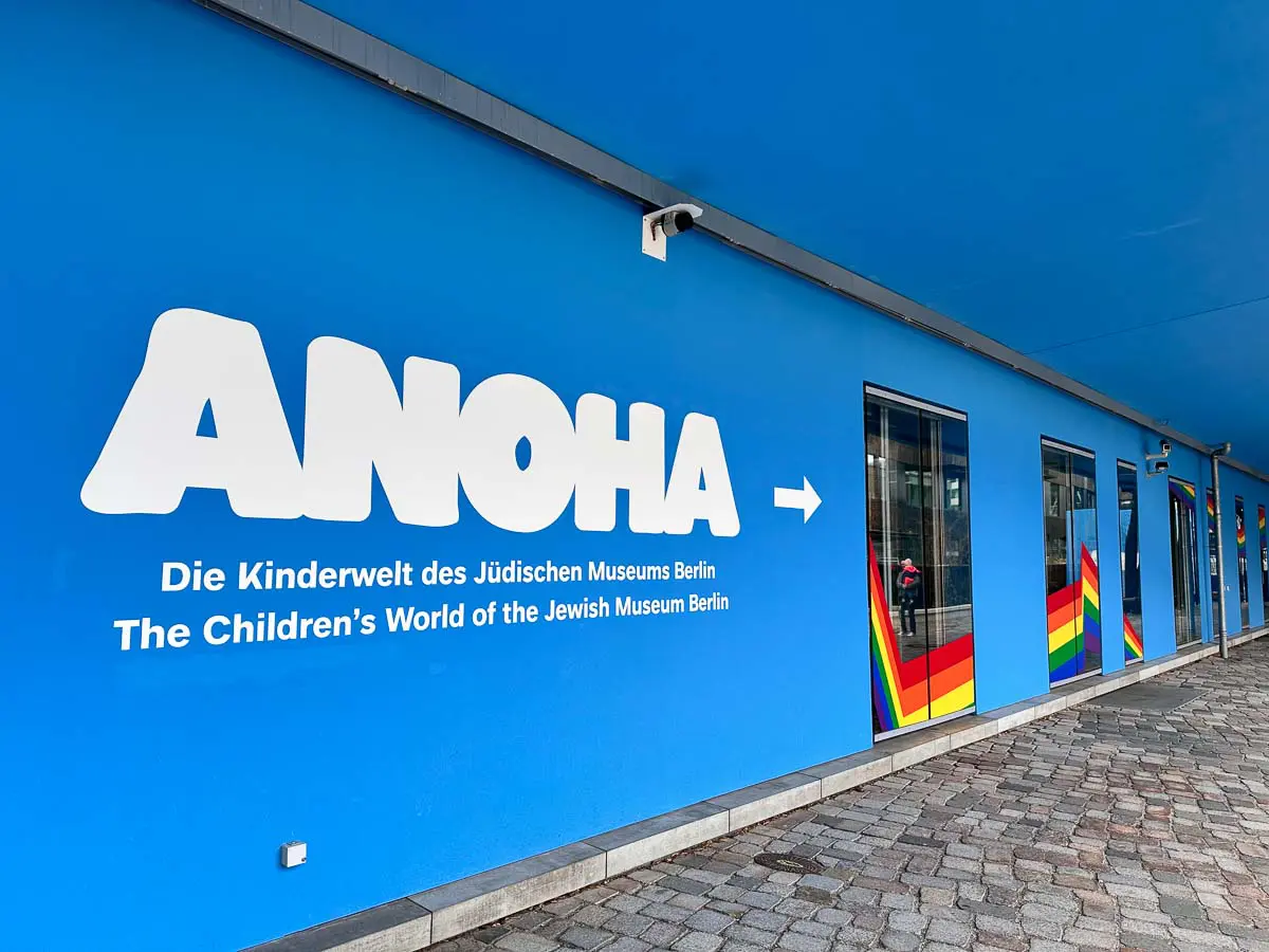 Blaue Außenwand eines Gebäudes mit großer weißer Schrift "ANOHA" und einem Pfeil, der auf Deutsch und Englisch für die Kinderwelt des Jüdischen Museums Berlin wirbt - der perfekte Ort für einen Besuch mit Kindern in Berlin bei Regen. Regenbogengestreifte Fenster bringen Farbe ins Spiel.