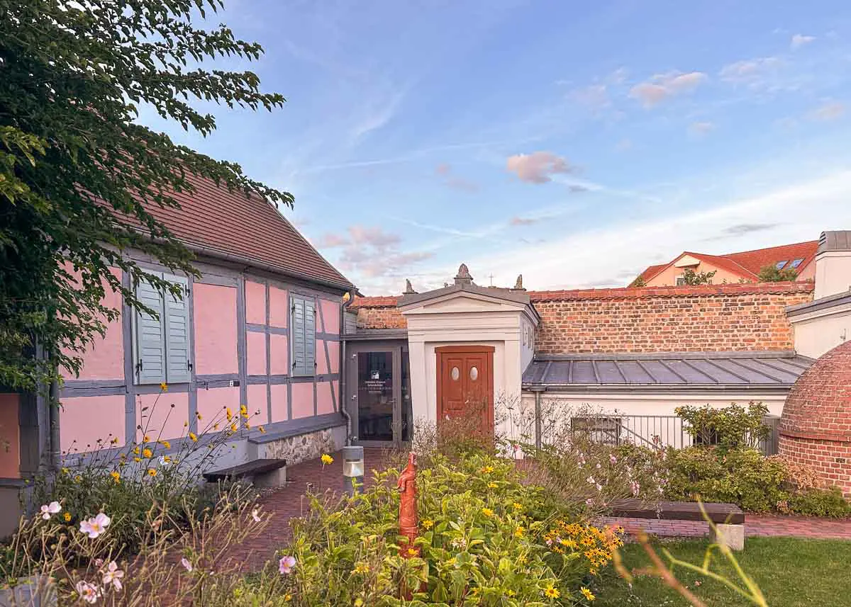 Ein bezauberndes rosa Fachwerkhaus mit blauen Fensterläden steht neben einem Backsteingebäude, umgeben von einem blühenden Garten unter einem teilweise bewölkten Himmel in der malerischen Region des Nationalparks Unteres Odertal.