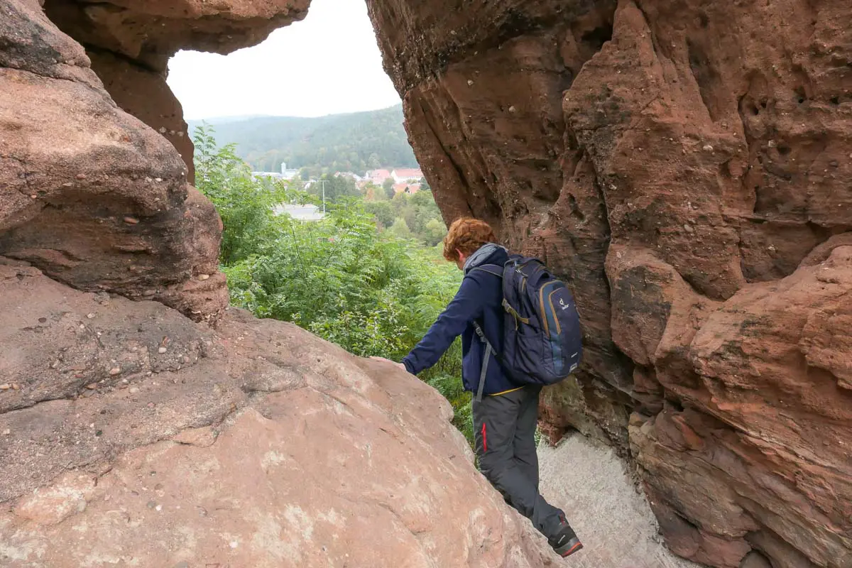 Eine Person mit blauer Jacke und Rucksack navigiert durch eine enge Felspassage im Pfälzerwald. Hinter der Öffnung locken üppiges Grün und ferne Hügel – ein unvergessliches Wandererlebnis.