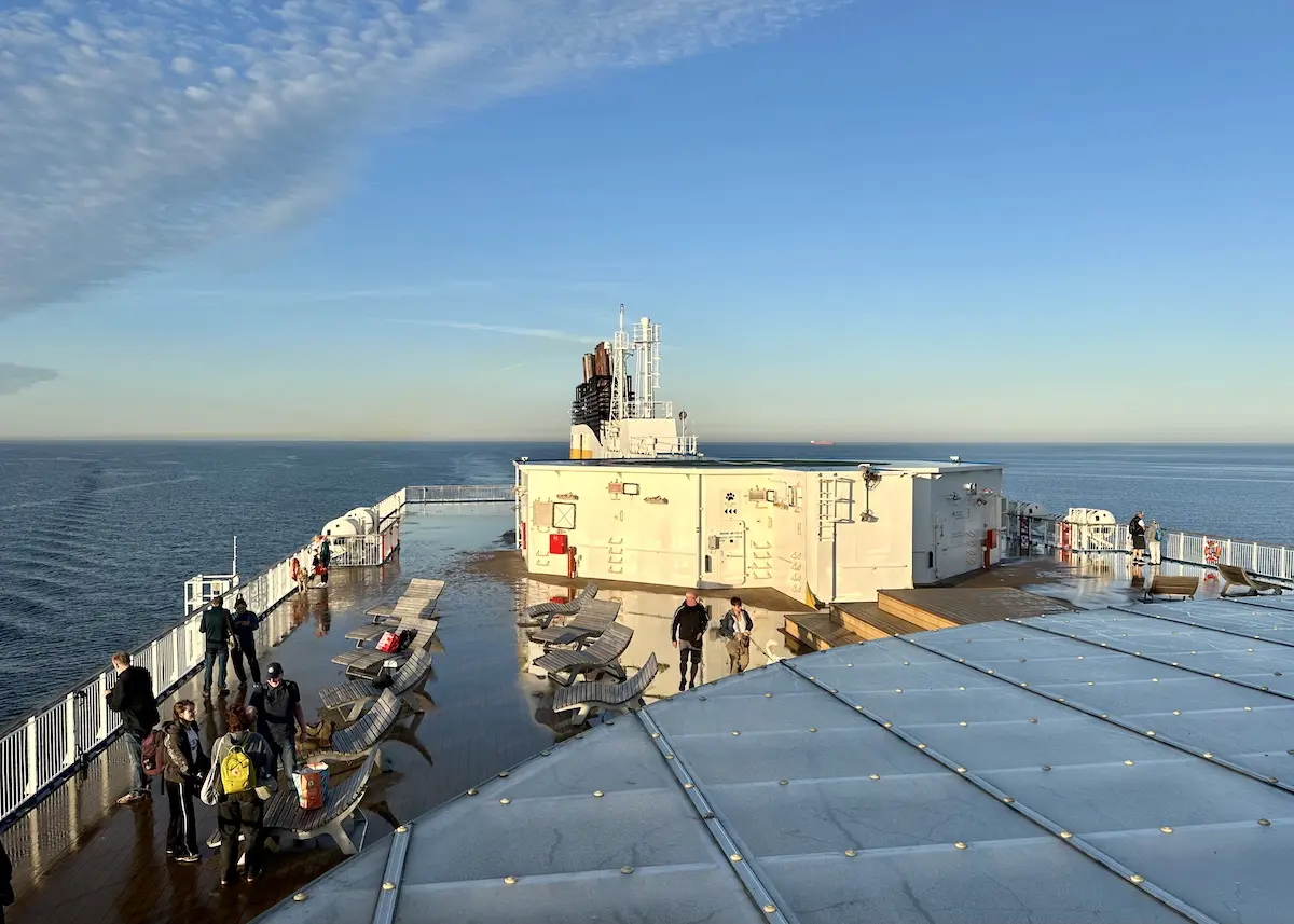 Unter strahlend blauem Himmel stehen und spazieren Menschen auf dem Oberdeck einer Fähre, von wo aus man in alle Richtungen Bänke, ein weißes Bauwerk und das Meer sehen kann. Die Sonne wirft lange Schatten über das Deck, während sich die Reisenden darauf vorbereiten, nach Norwegen zu reisen, und die bevorstehende malerische Reise in vollen Zügen genießen.
